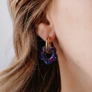 Créoles Oia Azuria dorées avec fleur en résine violette posées sur fond clair